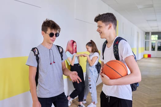 Group of teenage students talking standing outside school building, two males with backpacks basketball ball in focus, schoolgirls in corridor background. Lifestyle, communication, youth concept