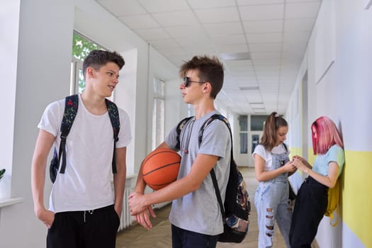 Group of teenage students talking standing outside school building, two males with backpacks basketball ball in focus, schoolgirls in corridor background. Lifestyle, communication, youth concept