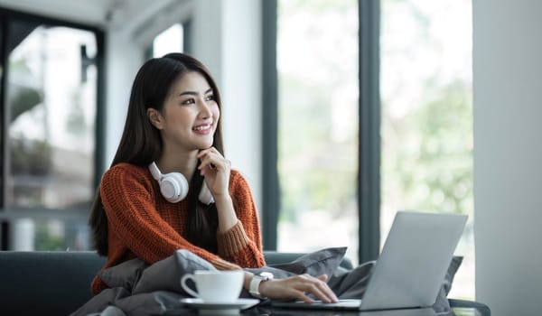 asian young woman listening music with headphone and streaming music from laptop on sofa relaxing at home..