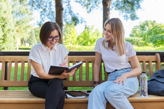 Woman teacher and girl student teenager sitting on the bench in park talking, testing, learning