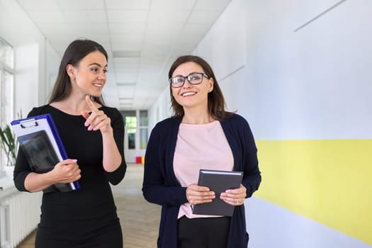 Two colleagues of businesswoman, teacher walking and talking on corridor of office, school. Positive smiling young and middle aged females, business education professions office workers concept