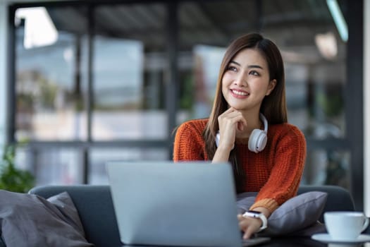 asian young woman listening music with headphone and streaming music from laptop on sofa relaxing at home..