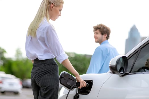 Focus hand insert EV charger to electric vehicle at public charging point in car park with blur business people in backdrop, eco-friendly lifestyle by rechargeable car for progressive concept.