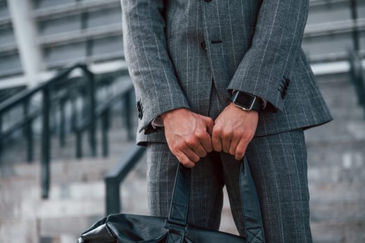 Holds bag in hands. Young businessman in grey formal wear is outdoors in the city.