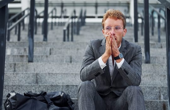 Smoking cigarette. Young businessman in grey formal wear is outdoors in the city.