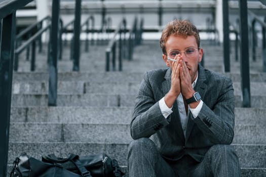 Smoking cigarette. Young businessman in grey formal wear is outdoors in the city.