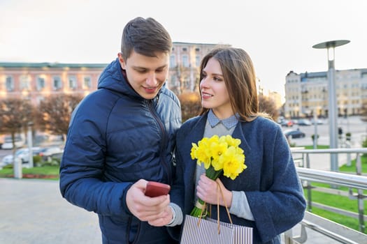 Young couple in love on Valentine's day walking in city, happy girl with bouquet of flowers and smiling guy looking together into smartphone hugging