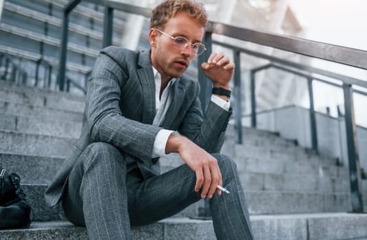 Smoking cigarette. Young businessman in grey formal wear is outdoors in the city.