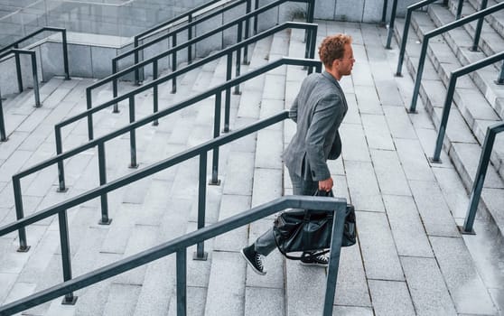 Holds bag in hands. Young businessman in grey formal wear is outdoors in the city.