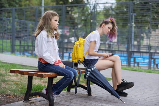 Children, city, lifestyle. Two girls sitting on bench in city park, near sports court, children look to the side