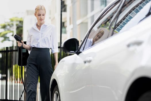 Progressive businesswoman insert charger plug from charging station to her electric vehicle with apartment condo building in background. Eco friendly rechargeable car powered by sustainable energy.