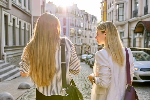 Two young women with laptop bags walking along street of sunset city, back view. Urban style background, female university students, colleagues office workers women