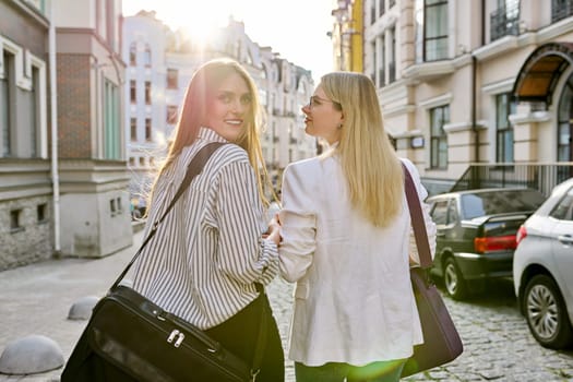 Two young women with laptop bags walking along street of sunset city, back view. Urban style background, female university students, colleagues office workers women