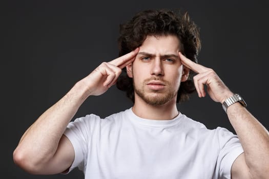 young bearded man in casual white t-shirt pointing to head with both hands finger on dark gray background. great idea