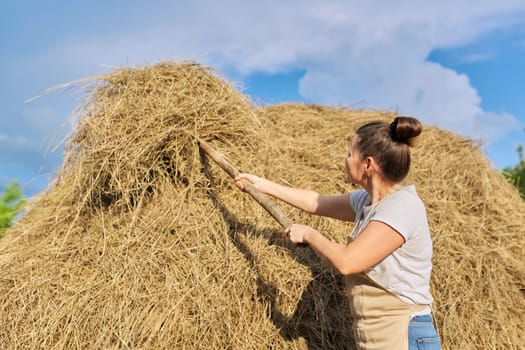 Woman farmer working with dry grass, straw. Haystack, countryside blue sky background