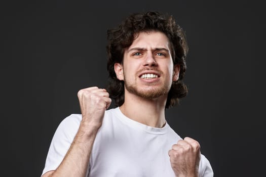 very excited young bearded man showing victory gesture holding fists raising hands up on gray background