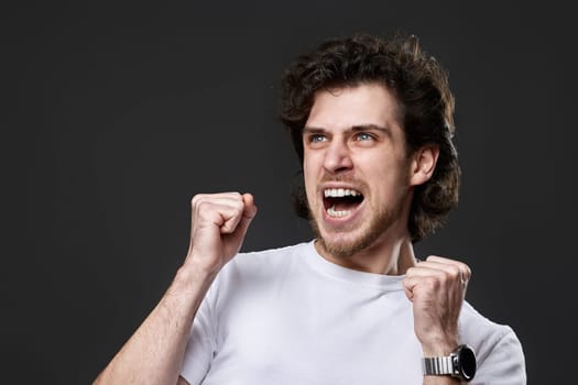 very excited young bearded man showing victory gesture holding fists raising hands up on gray background