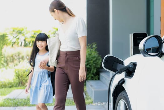 Focus electric vehicle recharging at home charging station plugged in with EV charger device with blurred background of progressive mother and daughter walking as concept for sustainability of energy.