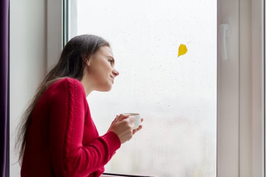 Portrait of young woman looking out the window with raindrops, autumn yellow leaf. Female enjoying a cup of coffee