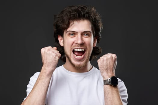 very excited young bearded man showing victory gesture holding fists raising hands up on gray background