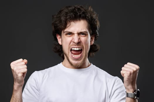 very excited young bearded man showing victory gesture holding fists raising hands up on gray background