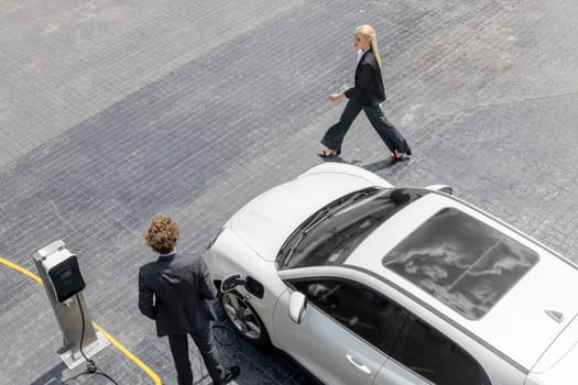 Aerial view progressive lifestyle of businesspeople with electric car connected to charging station at city center public parking car. Eco friendly rechargeable car powered by alternative clean energy