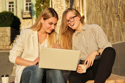 Two young beautiful women having fun looking into the laptop monitor, business females sitting on steps in city street, university students laughing resting