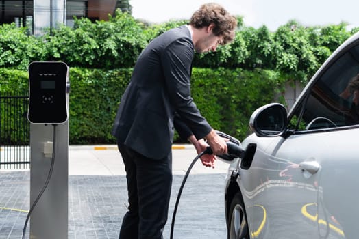 Progressive businessman with electric car recharging at public charging station at modern city residential area. Eco friendly rechargeable car powered by alternative clean energy.