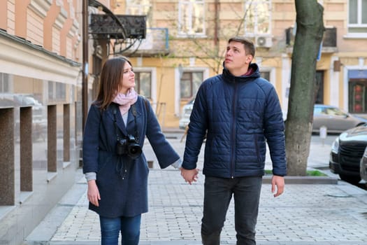 Outdoor portrait of happy couple in love walking along street of winter spring city holding hands, woman with camera