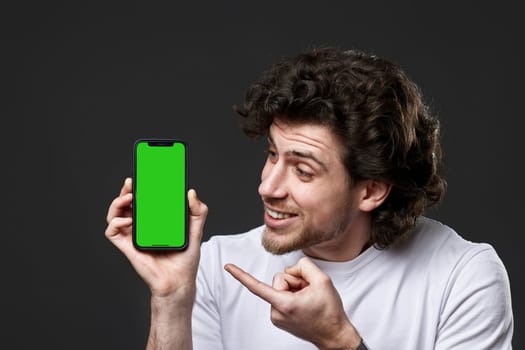 young bearded curly man pointing finger at green screen of phone on dark gray background