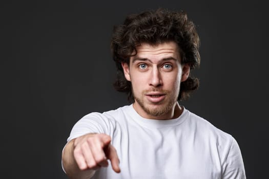 surprised caucasian man pointing finger at you on dark gray background. guy asks a question - is it you