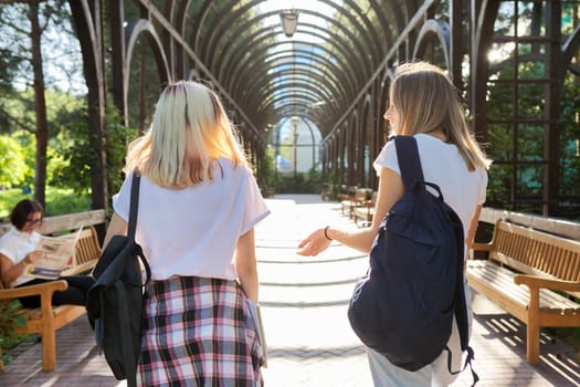 Two happy smiling talking girls teenagers students walking together, young women with backpacks, sunny day in the park background, back view