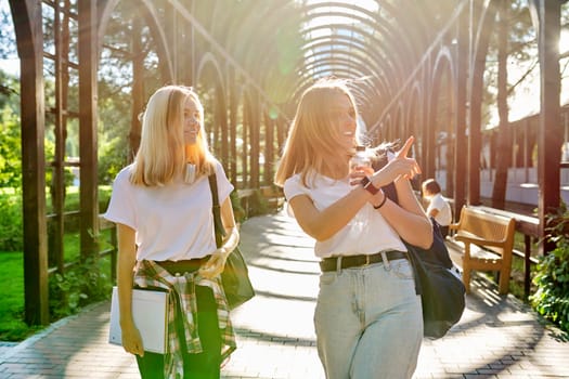 Two happy smiling talking girls teenagers students walking together, young women with backpacks, sunny day in the park background