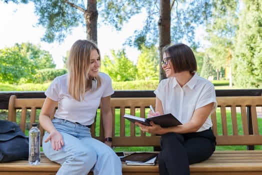 Woman social worker talking to girl teenager writing interviews in notebook, women sitting on bench in park