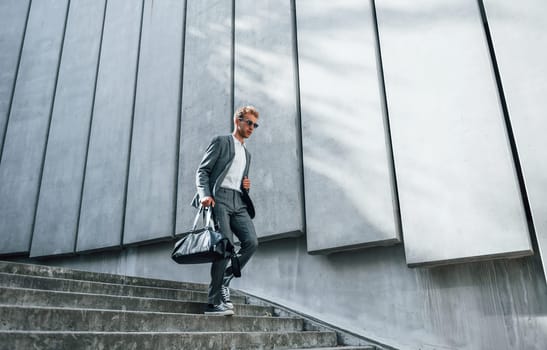 Going down by the stairs. Young businessman in grey formal wear is outdoors in the city.