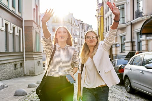 Two young beautiful happy women university students walking along city street, women smiling laughing looking ahead waving hand