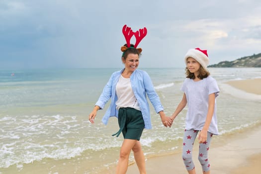Happy mom and daughter child in Santa hat walking holding hands along the beach. Family celebrating Christmas and New Year at tropical resort, copy space, vacations travel tourism concept