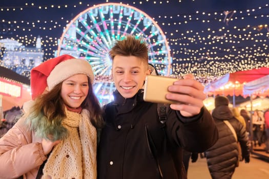 Christmas time, New Year holidays. Young people, couple of teenagers having fun at Christmas market, taking selfie photo on smartphone, talking laughing, garlands of evening city, Ferris wheel