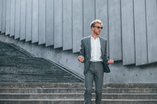 Posing for a camera. Young businessman in grey formal wear is outdoors in the city.