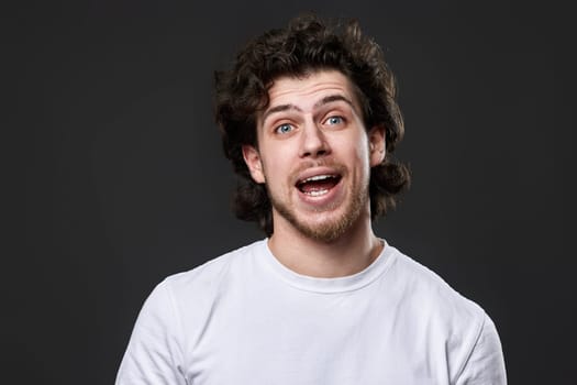 portrait of young smiling curly man in white t-shirt on dark gray background