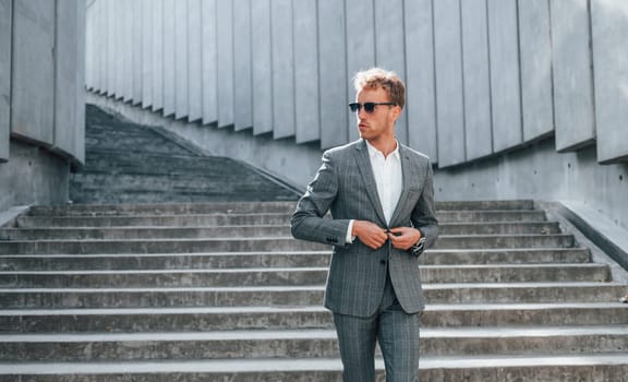 Posing for a camera. Young businessman in grey formal wear is outdoors in the city.