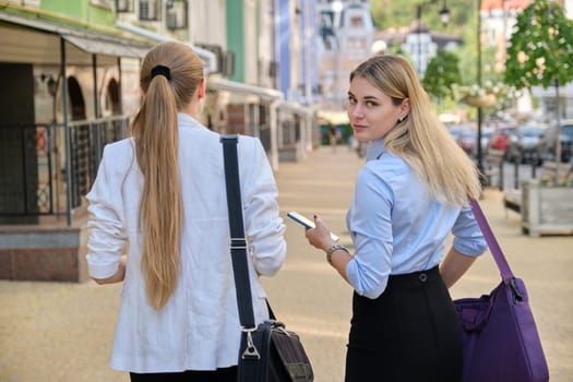 Back shot of two young business women walking and talking in the city, female office employees