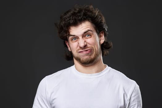 portrait of young funny curly man in white t-shirt grimacing on dark gray background
