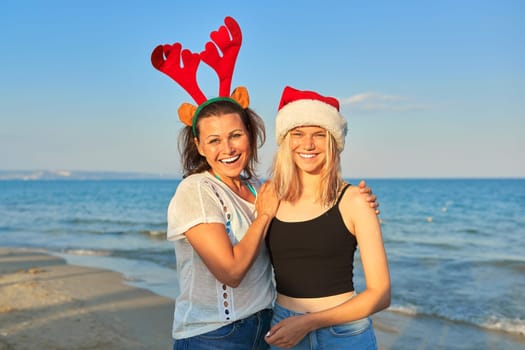 Happy smiling mother and teenage daughter in Santa Claus hat on the beach, New Year and Christmas holidays together with family. Travel, vacation, sea ocean, people concept