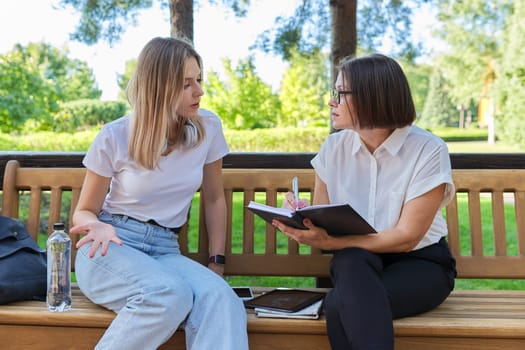 Woman teacher and girl student teenager sitting on the bench in park talking, testing, learning