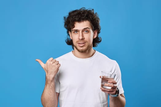 handsome bearded man in white t-shirt with drinking glass of water pointing to the side on blue background