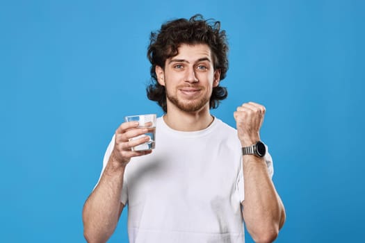 handsome curly man in white t-shirt with raised arm holding glass of water smiling happy on blue background