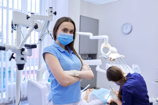 Portrait of confident female dentist in medical mask with crossed arms, professional doctor looking at camera in dental clinic