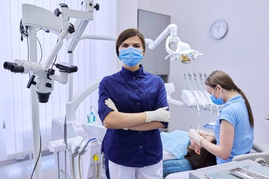 Portrait of young female doctor dentist in protective medical face mask with arms crossed in dental office, treating woman patient in chair background. Medicine, dentistry and health care concept