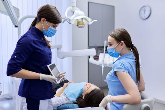 Dentistry, woman patient in chair in dental office, doctor colleagues consult looking in digital tablet. Medicine, dentistry and health care concept